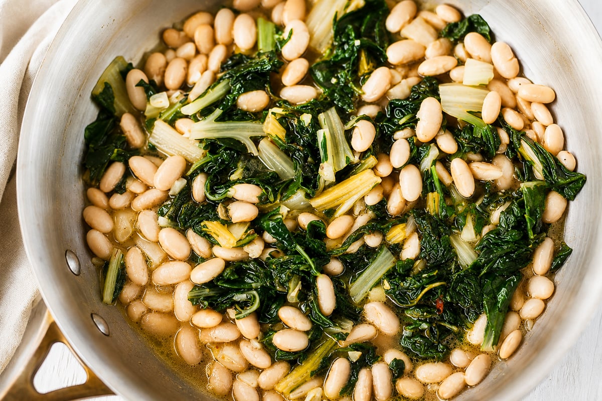 White beans and cooked greens, likely rainbow chard or spinach, are simmering in a light broth in a stainless steel pan. The dish appears warm, healthy, and freshly prepared.