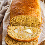 A loaf of Anadama Bread on parchment paper with two slices cut, one slice topped with melting butter, all on a wooden surface.