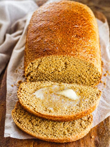 A loaf of Anadama Bread on parchment paper with two slices cut, one slice topped with melting butter, all on a wooden surface.