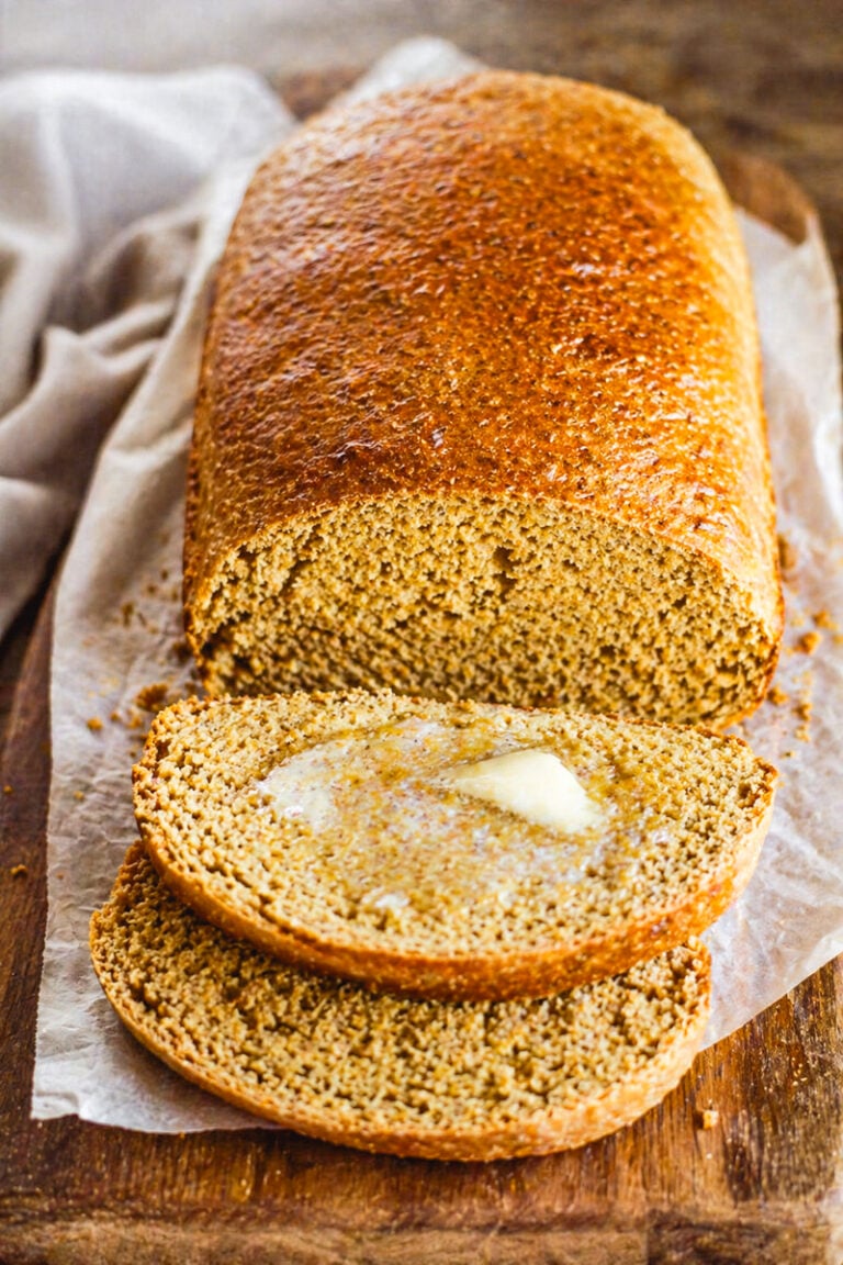 A loaf of Anadama Bread on parchment paper with two slices cut, one slice topped with melting butter, all on a wooden surface.