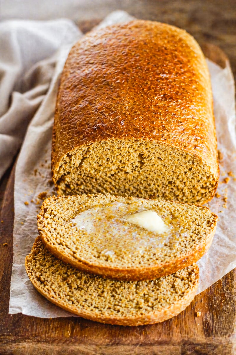 A loaf of Anadama Bread on parchment paper with two slices cut, one slice topped with melting butter, all on a wooden surface.