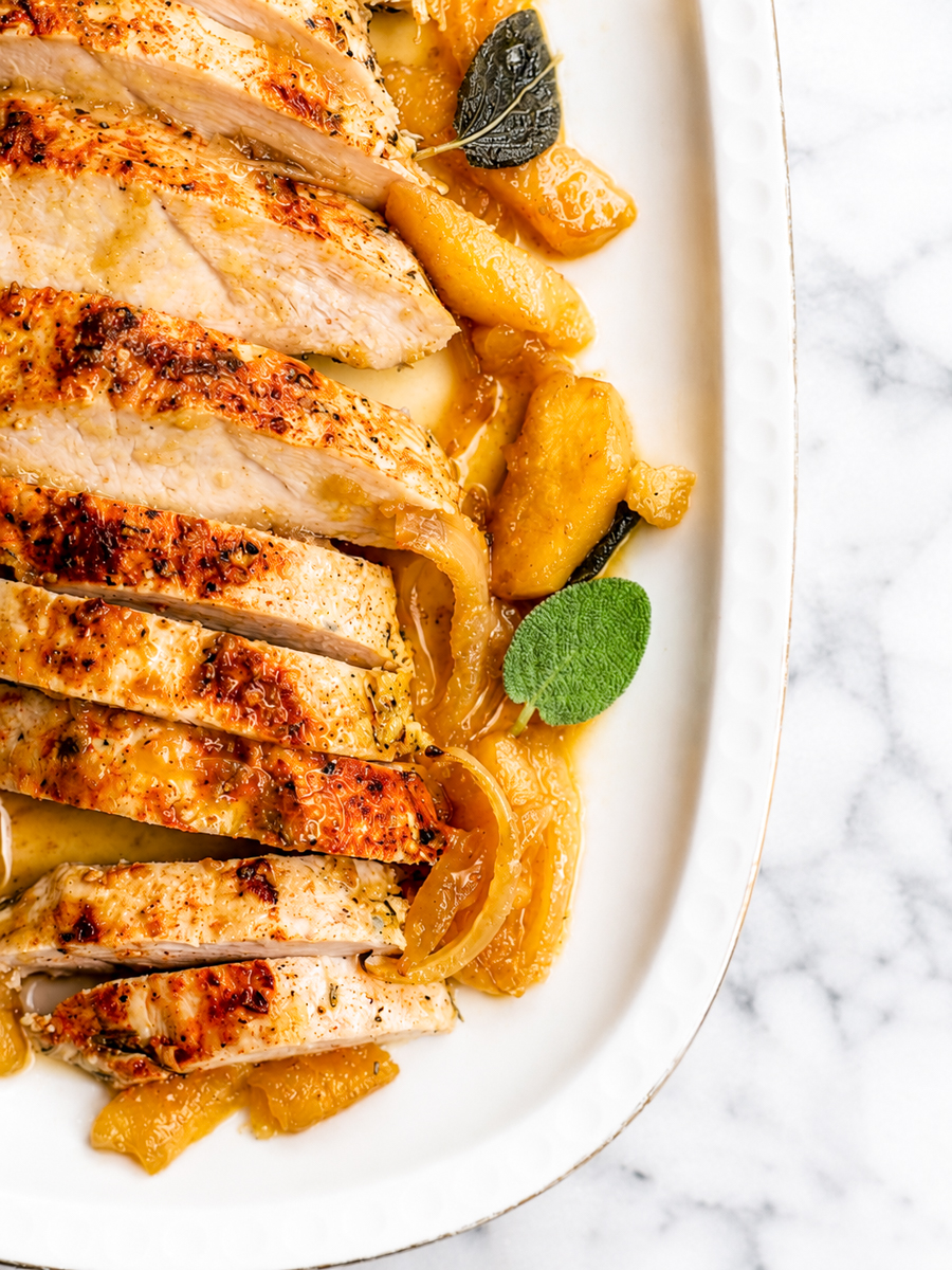 Sliced roasted chicken breast arranged on a white platter, garnished with caramelized apples and fresh sage leaves, sits alongside tender baked turkey tenderloin on a marble surface.