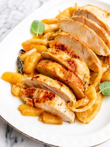 Sliced roasted turkey breast, reminiscent of a succulent Baked Turkey Tenderloin, served on a white plate and garnished with cooked apples, onions, and fresh sage leaves on a marble background.