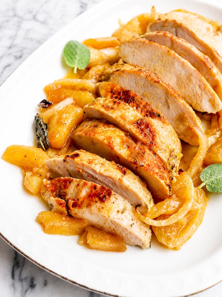 Sliced roasted turkey breast, reminiscent of a succulent Baked Turkey Tenderloin, served on a white plate and garnished with cooked apples, onions, and fresh sage leaves on a marble background.