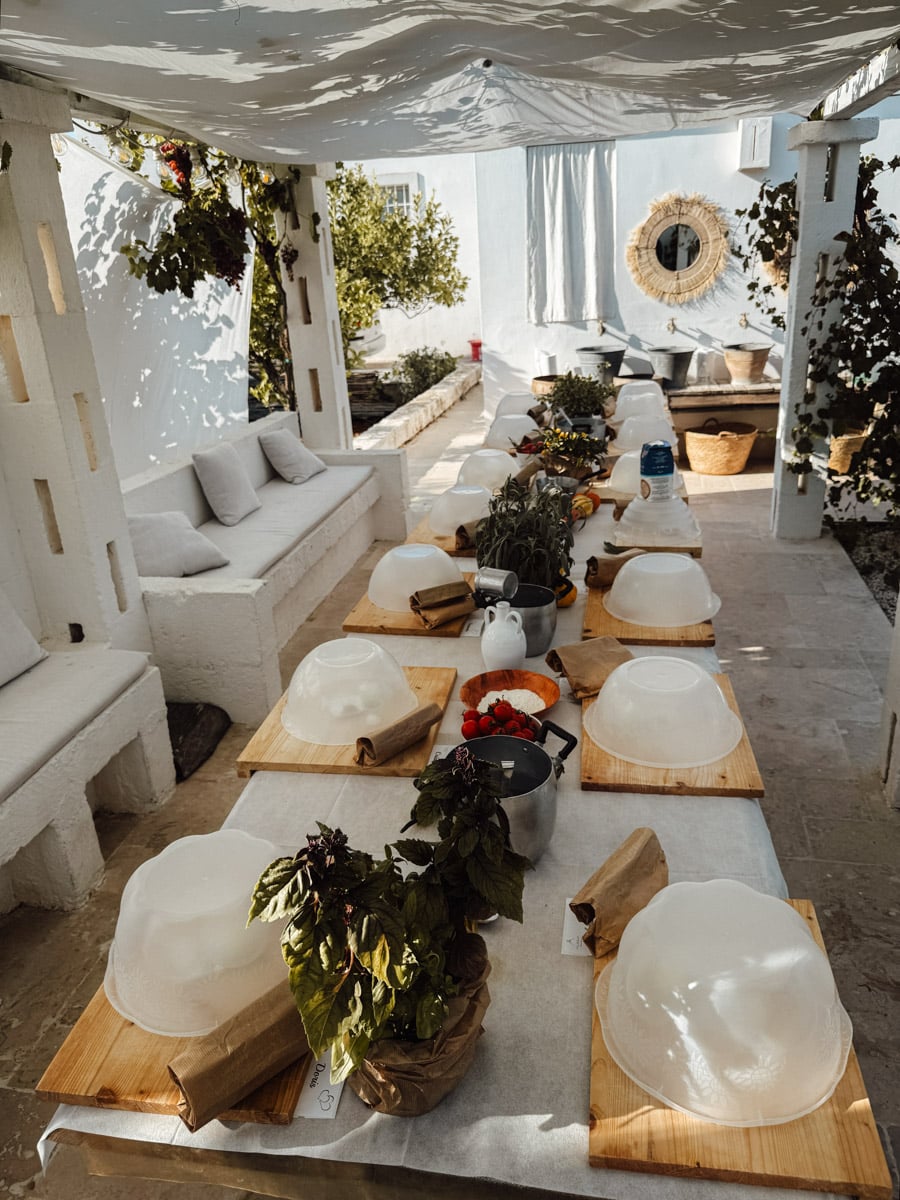 A long outdoor dining table is set for a meal with wooden boards, glass bowls, herbs, and vegetables. White cushions line built-in benches as sunlight filters through a canopy in a bright courtyard inspired by Ostuni, Italy.