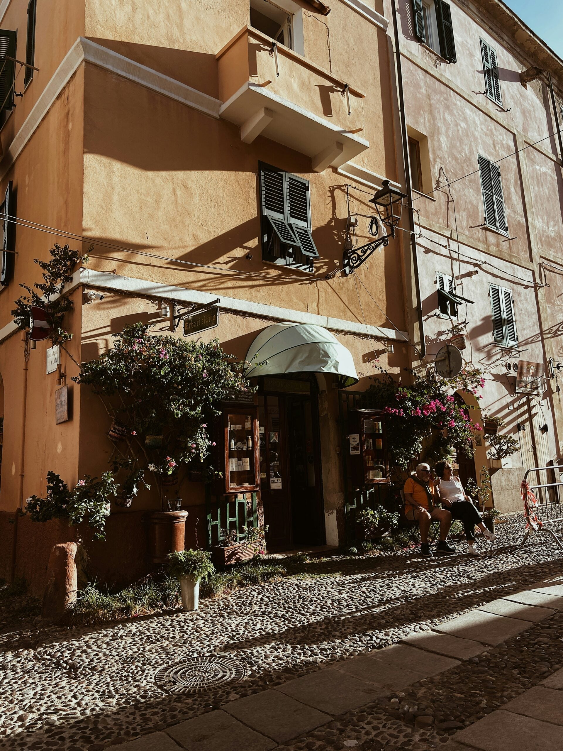 A cozy street scene in Sassari, Sardinia, shows two people sitting on a bench outside a sunlit, peach-colored building with green shutters, potted plants, flowers, and a cobblestone sidewalk.