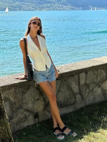 A woman wearing sunglasses, a white vest, and denim shorts leans against a stone wall by a lake—an effortless look for what to wear in Italy. Green hills, sailboats, and blue water stretch out behind her on this sunny day.