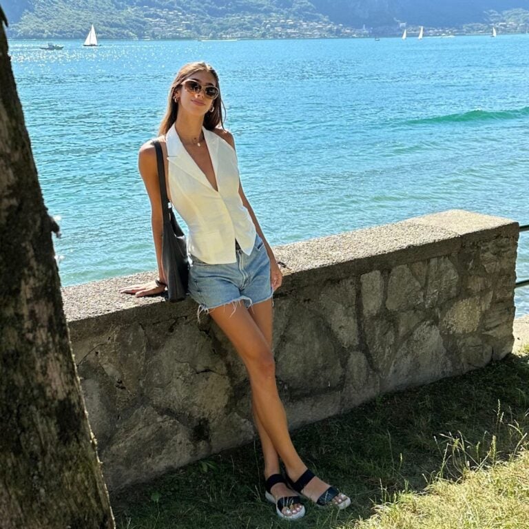 A woman wearing sunglasses, a white vest, and denim shorts leans against a stone wall by a lake—an effortless look for what to wear in Italy. Green hills, sailboats, and blue water stretch out behind her on this sunny day.