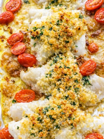 Baked Cod fillets topped with a golden, crispy breadcrumb crust, garnished with chopped herbs and surrounded by roasted cherry tomatoes on a baking tray.