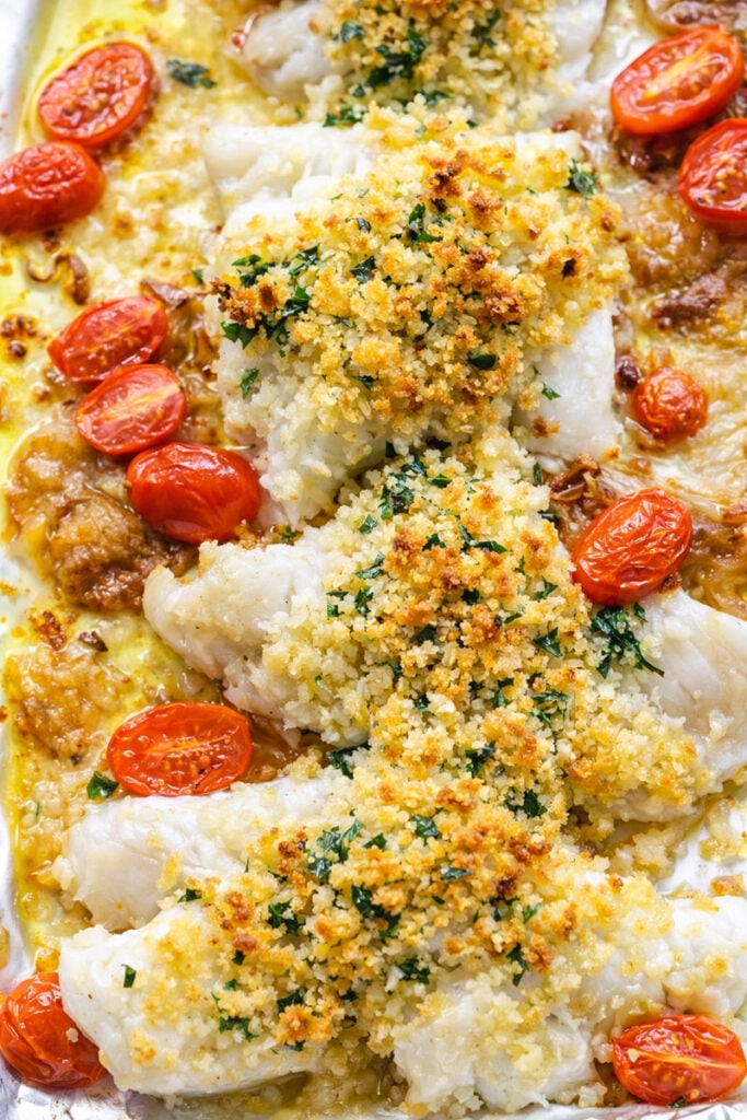 Baked Cod fillets topped with a golden, crispy breadcrumb crust, garnished with chopped herbs and surrounded by roasted cherry tomatoes on a baking tray.