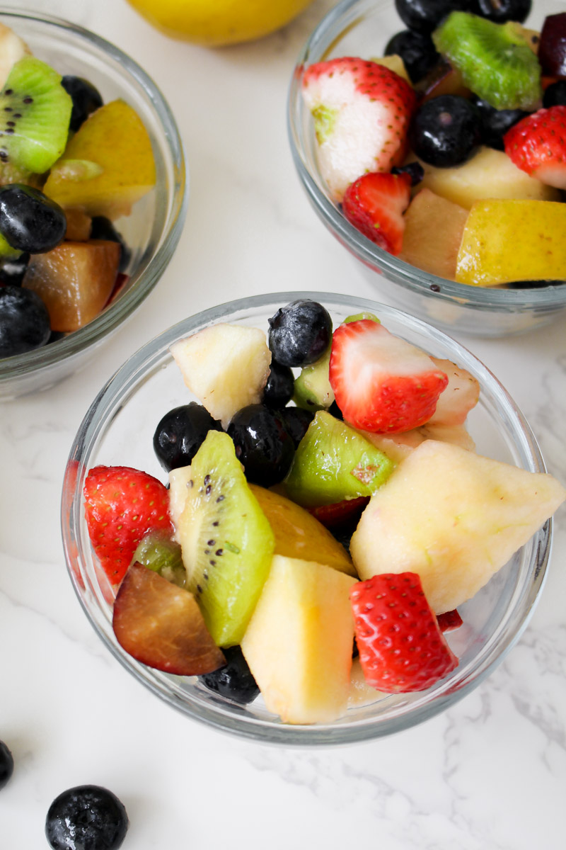 Italian fruit salad presented in a glass serving dish, combining chopped apples, bananas, kiwis, plums, strawberries, and blueberries.