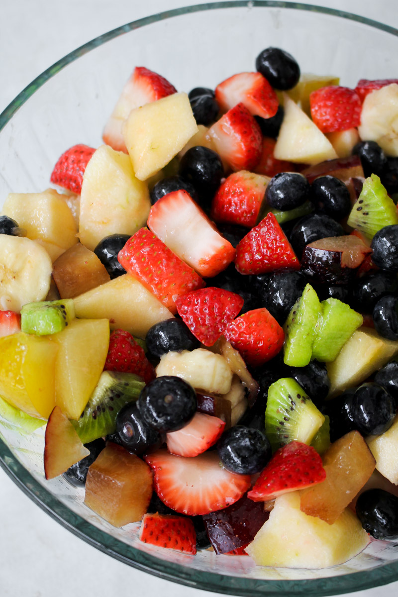 Italian fruit salad presented in a glass serving dish, combining chopped apples, bananas, kiwis, plums, strawberries, and blueberries.