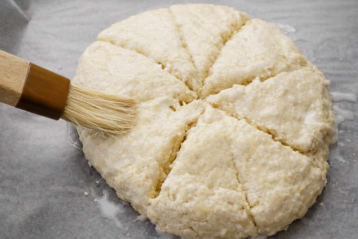 A round, unbaked Scottish Scones recipe dough divided into wedges sits on parchment paper as a pastry brush applies a liquid glaze to the surface.