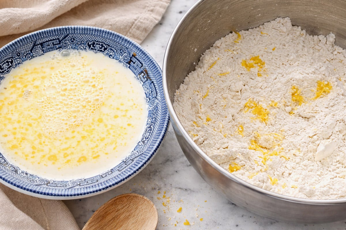A blue and white bowl with a beaten egg and milk mixture sits next to a metal bowl containing flour and bits of yellow butter, ready for a classic Scottish Scones recipe, with a wooden spoon nearby on the marble surface.