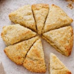 A round batch of golden-brown scones, made from a classic Scottish Scones recipe, is cut into eight triangular pieces on a lightly floured surface, with a bowl of butter in the background.