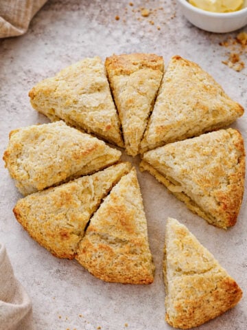 A round batch of golden-brown scones, made from a classic Scottish Scones recipe, is cut into eight triangular pieces on a lightly floured surface, with a bowl of butter in the background.