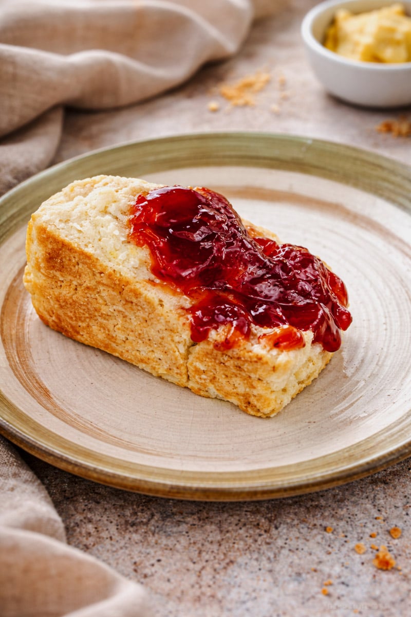 A golden biscuit made from a classic Scottish Scones recipe is topped with red strawberry jam on a beige plate, accompanied by a cloth napkin and a small bowl of butter in the background.