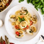 A plate of linguine alle vongole with clams, cherry tomatoes, and parsley sits on a white surface next to fresh parsley, dried red chili peppers, and a fork. A pan with more pasta and clams is partially visible.