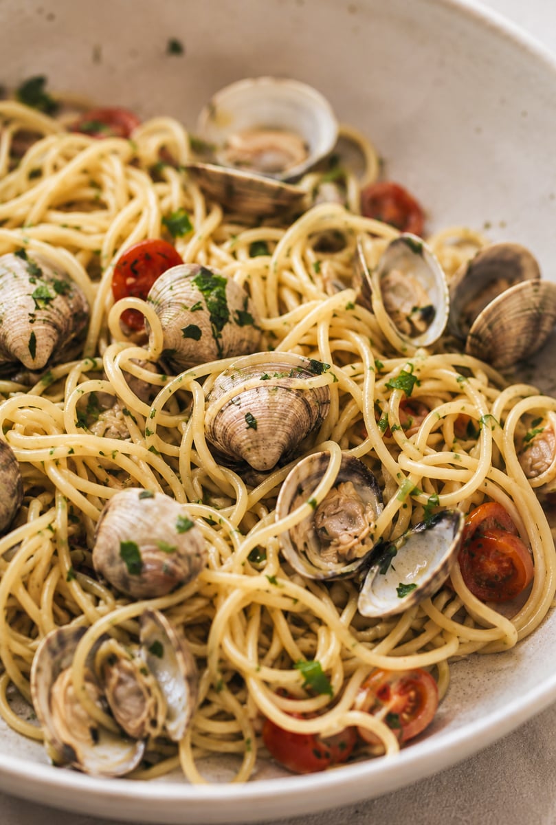A close-up of linguine alle vongole with clams, halved cherry tomatoes, and chopped parsley, served in a white bowl.