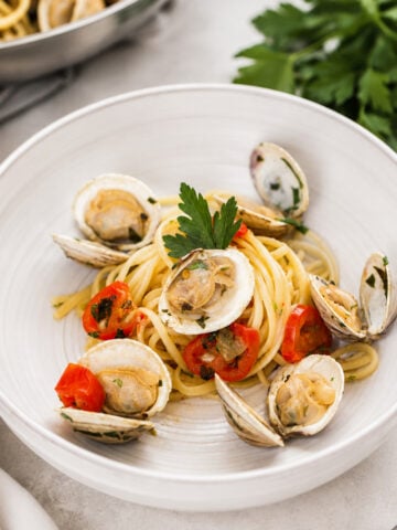 A white plate of linguine alle vongole is topped with clams, cherry tomatoes, fresh parsley, and olive oil, garnished with parsley, with a bunch of parsley in the background.