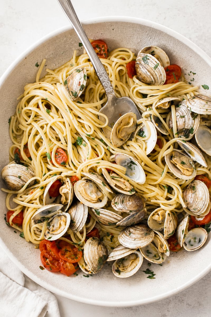 A bowl of linguine alle vongole with clams, halved cherry tomatoes, chopped parsley, and a light sauce, served with a metal serving spoon.