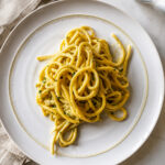A plate of creamy Bucatini alla Carbonara garnished with herbs is served on a white dish, placed on a light marble surface next to a beige cloth napkin and a glass of water.