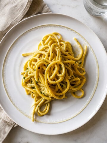 A plate of creamy Bucatini alla Carbonara garnished with herbs is served on a white dish, placed on a light marble surface next to a beige cloth napkin and a glass of water.