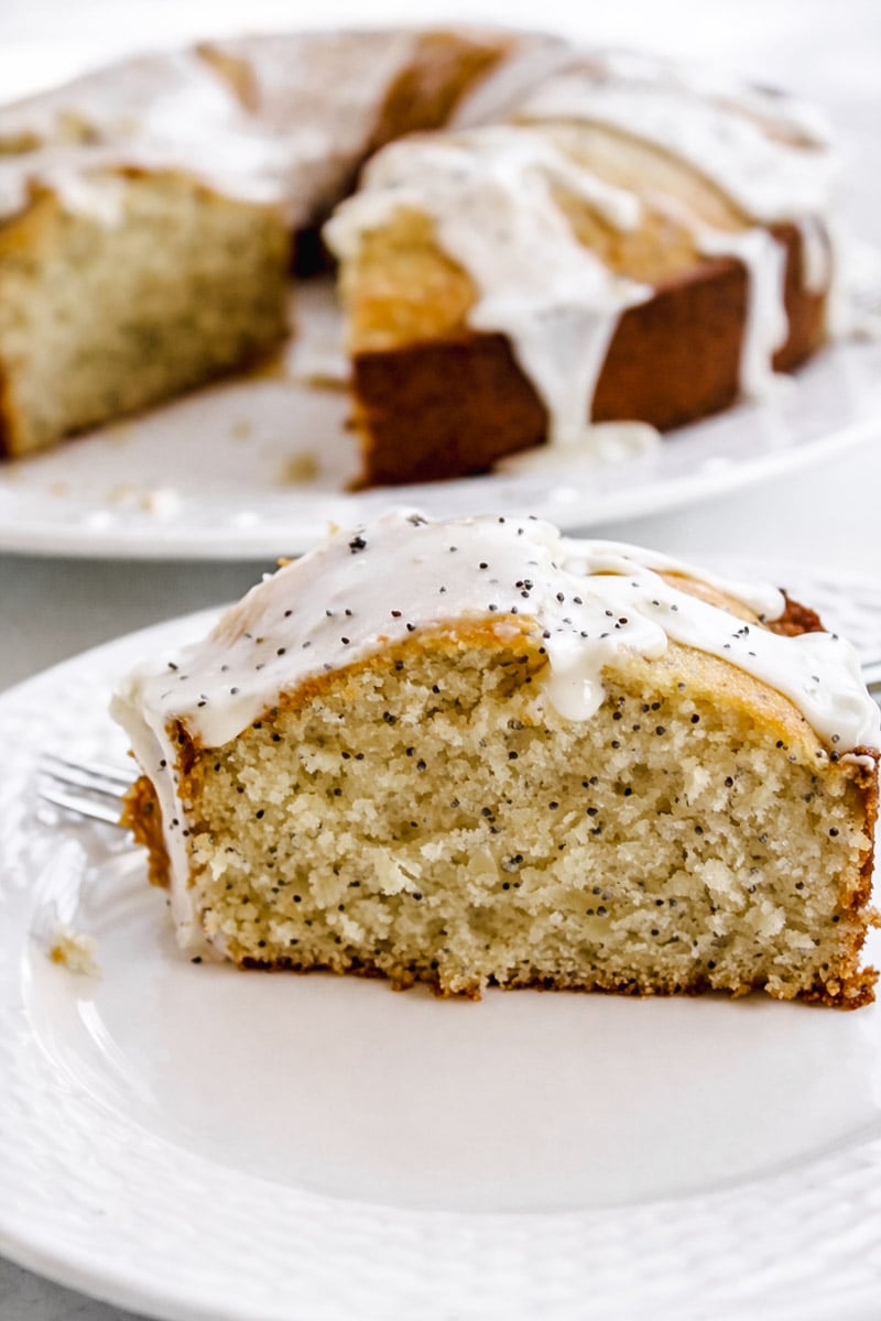 A close-up of a slice of Orange Poppyseed Cake with white icing, served on a white plate. The rest of the cake, also topped with icing, is visible in the background with a missing slice.