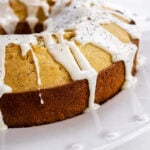An Orange Poppyseed Cake with a golden-brown crust, topped with white icing that drips down the sides, sits on a white plate.