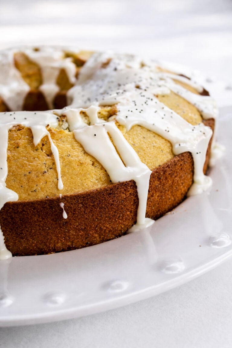 An Orange Poppyseed Cake with a golden-brown crust, topped with white icing that drips down the sides, sits on a white plate.