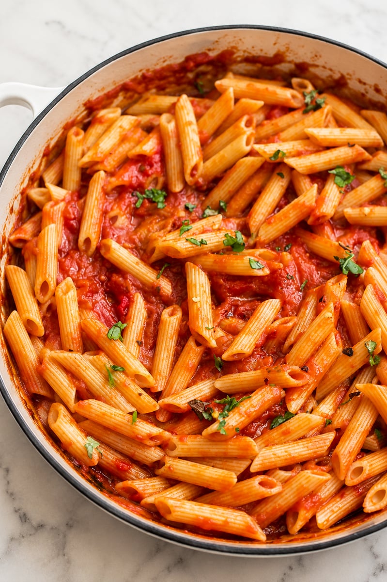 A white pan filled with penne pasta tossed in a vibrant red arrabbiata sauce, garnished with small pieces of chopped fresh herbs, sits on a marble surface.