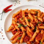 A white plate of penne arrabbiata with red tomato sauce and chopped herbs sits on a marble surface. Nearby are two red chili peppers, a garlic clove, and a glass bowl of red pepper flakes.