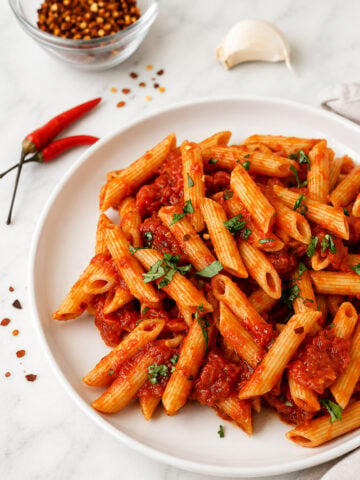 A white plate of penne arrabbiata with red tomato sauce and chopped herbs sits on a marble surface. Nearby are two red chili peppers, a garlic clove, and a glass bowl of red pepper flakes.