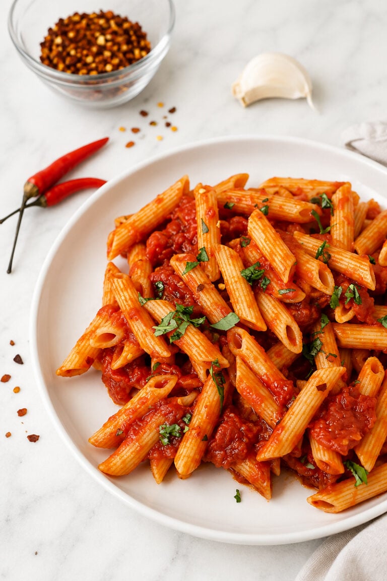 A white plate of penne arrabbiata with red tomato sauce and chopped herbs sits on a marble surface. Nearby are two red chili peppers, a garlic clove, and a glass bowl of red pepper flakes.