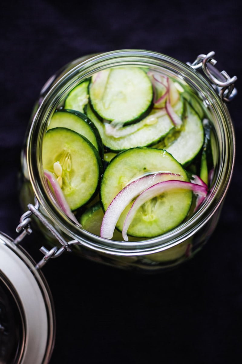 A glass jar of Easy Pickled Cucumbers and thinly sliced red onions, shown from above against a dark background. The open jar lid is visible at the edge, highlighting this simple, homemade treat.