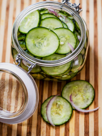 A glass jar of easy pickled cucumbers and red onions sits open on a striped wooden surface, with a few cucumber and onion slices scattered beside it.