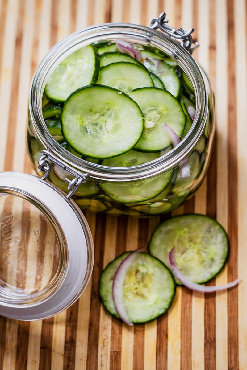 A glass jar of easy pickled cucumbers and red onions sits open on a striped wooden surface, with a few cucumber and onion slices scattered beside it.