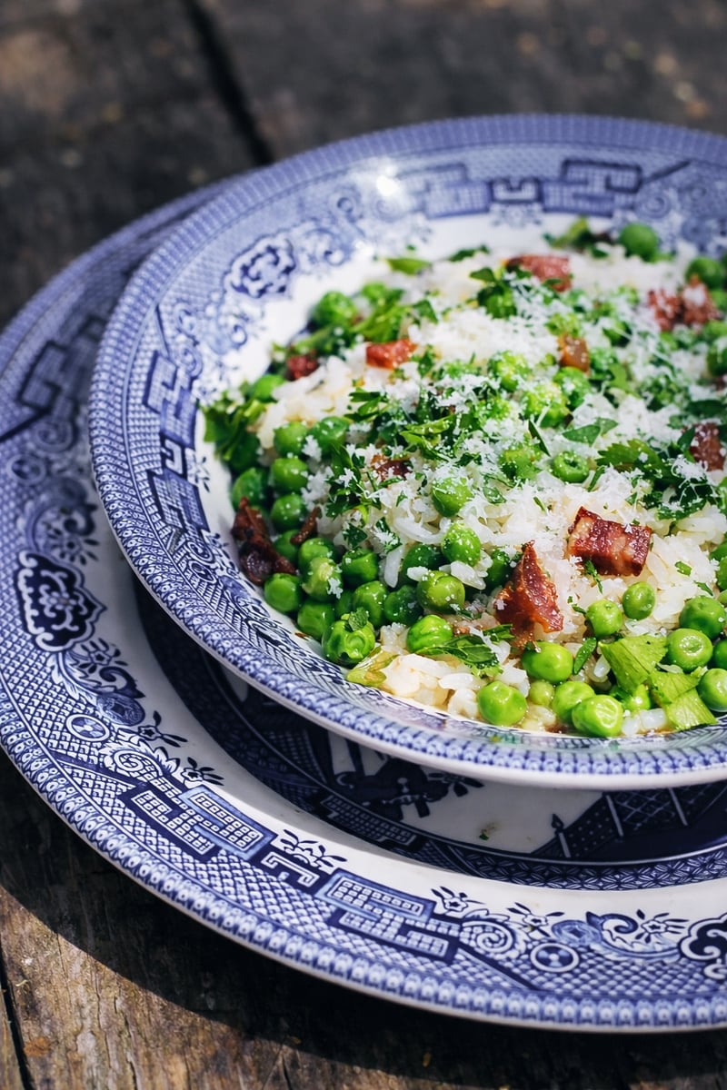 A blue and white patterned bowl holds Risi e Bisi-Rice and Peas with chopped herbs, grated cheese, and bacon, set on a matching plate atop a rustic wooden surface.