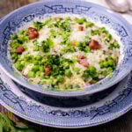 A blue and white patterned bowl filled with Risi e Bisi-Rice and Peas risotto, green peas, chopped parsley, grated cheese, and bacon sits on a matching plate atop a wooden table. A spoon and parsley garnish are nearby.
