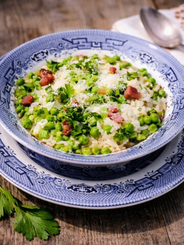 A blue and white patterned bowl filled with Risi e Bisi-Rice and Peas risotto, green peas, chopped parsley, grated cheese, and bacon sits on a matching plate atop a wooden table. A spoon and parsley garnish are nearby.