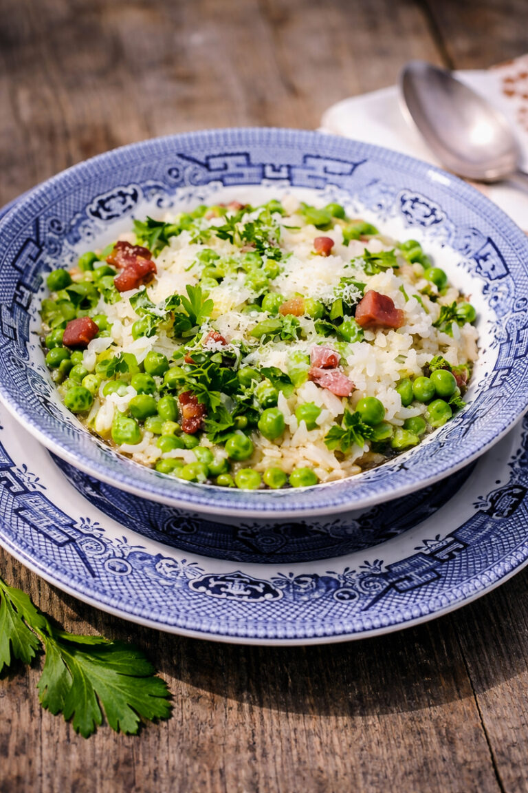 A blue and white patterned bowl filled with Risi e Bisi-Rice and Peas risotto, green peas, chopped parsley, grated cheese, and bacon sits on a matching plate atop a wooden table. A spoon and parsley garnish are nearby.