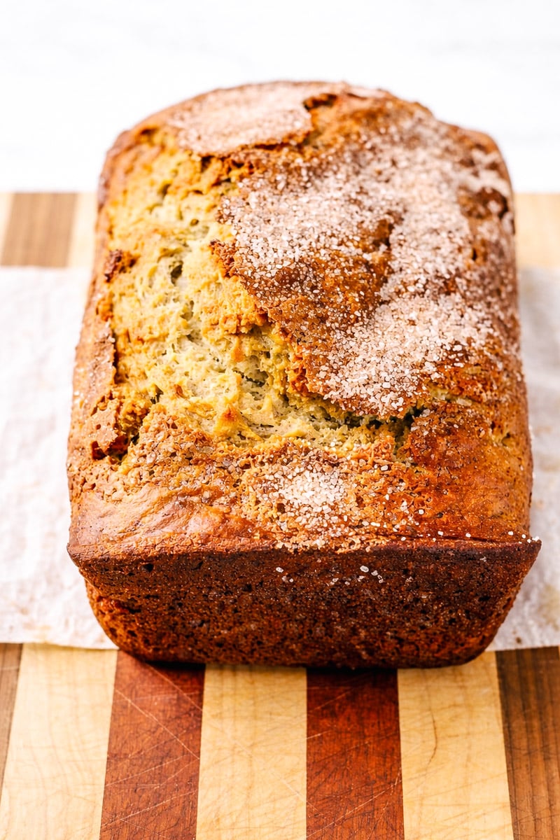 A golden-brown loaf of sourdough banana bread topped with a sprinkle of sugar sits on a wooden cutting board with alternating light and dark stripes.