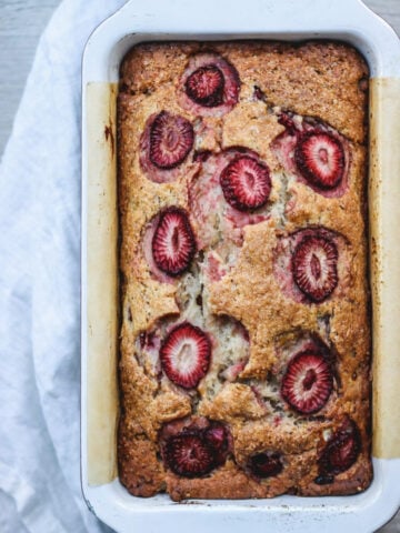 A loaf of strawberry banana bread baked in a white rectangular pan, with sliced strawberries visible on top and a golden-brown crust. The pan is resting on a light-colored cloth.