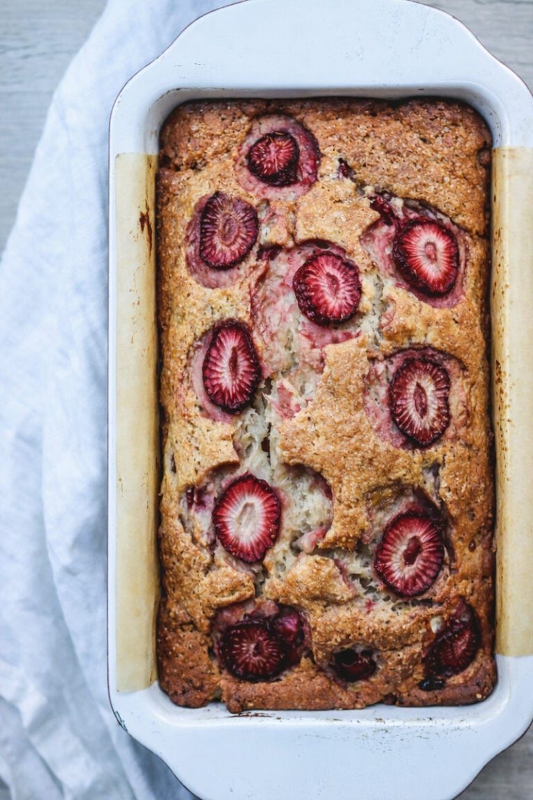 A loaf of strawberry banana bread baked in a white rectangular pan, with sliced strawberries visible on top and a golden-brown crust. The pan is resting on a light-colored cloth.