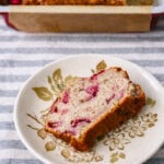 A slice of strawberry banana bread with visible strawberry pieces sits on a decorative plate, with the rest of the loaf in a baking dish in the background, all placed on a striped cloth.
