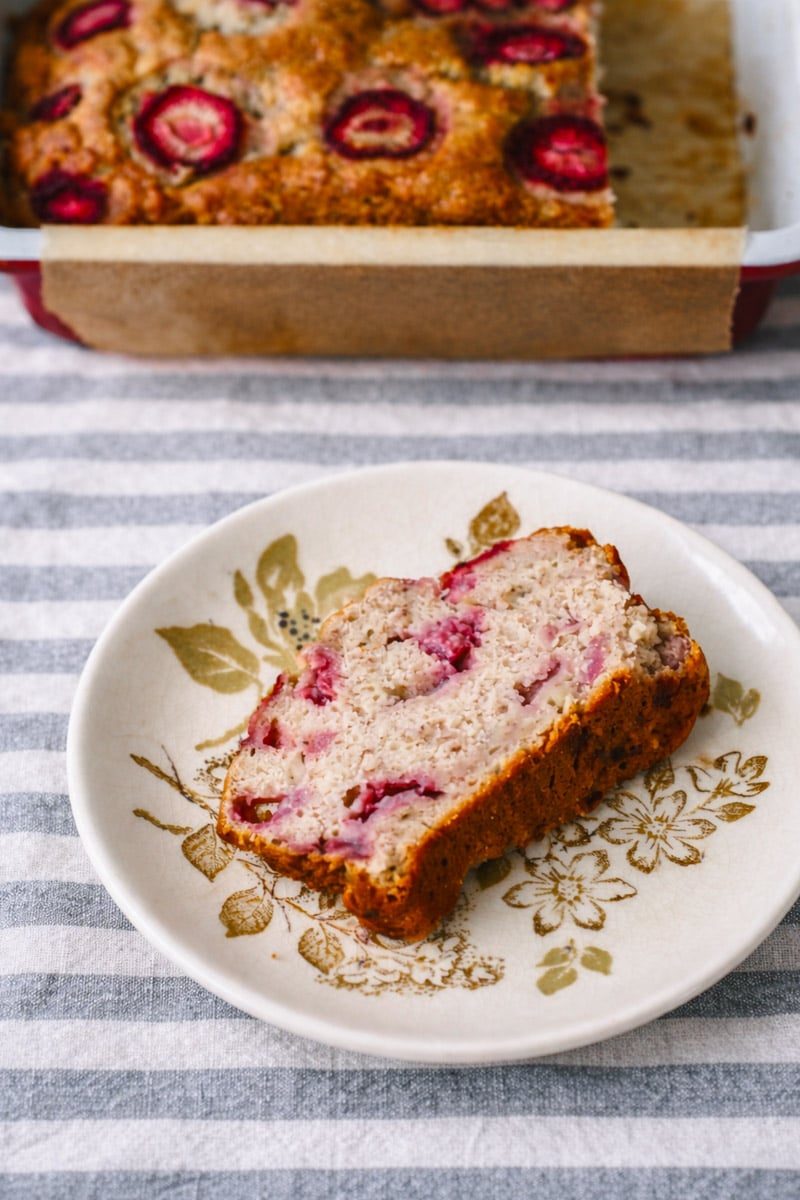 A slice of strawberry banana bread with visible strawberry pieces sits on a decorative plate, with the rest of the loaf in a baking dish in the background, all placed on a striped cloth.