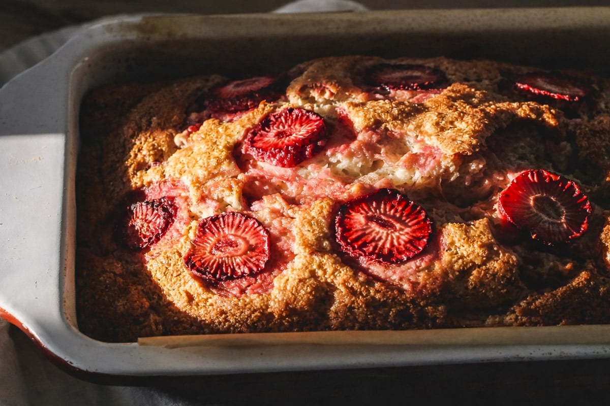 A close-up of freshly baked Strawberry Banana Bread in a rectangular dish, topped with slices of strawberries. The loaf is golden brown, with sunlight highlighting its textured surface and vibrant red strawberries.