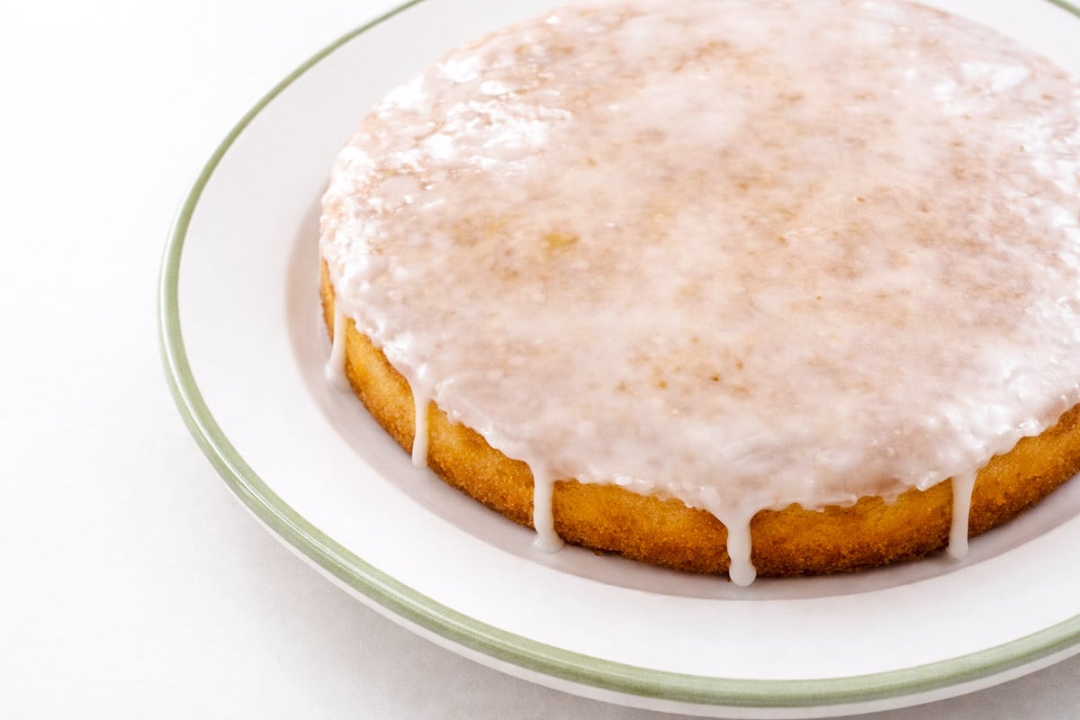 A round, plain lemon cake with white glaze icing dripping down the sides sits on a white plate with a green rim. The cake appears simple and homemade, with a smooth, shiny glaze on top.