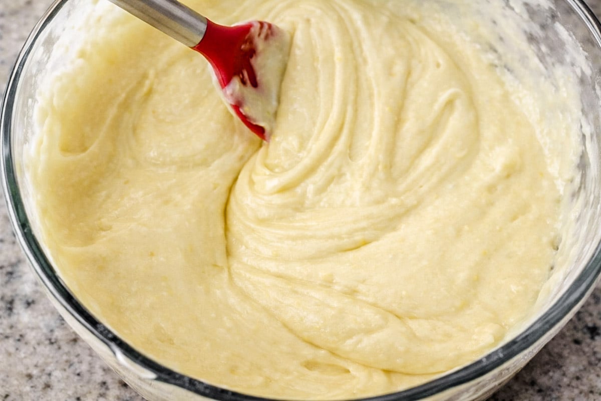 A glass bowl filled with creamy lemon cake batter being mixed with a red silicone spatula on a speckled countertop.