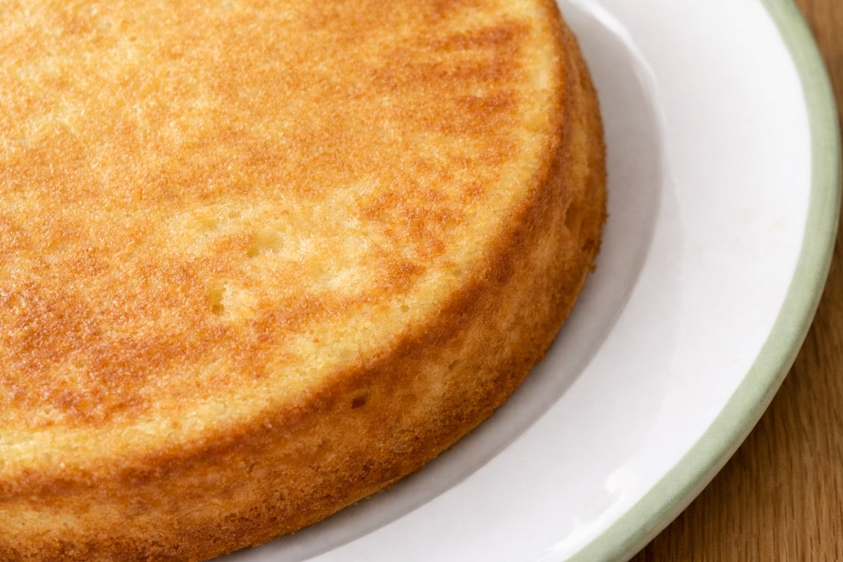 A close-up of a plain, golden-brown lemon cake on a white plate with a green rim, sitting on a wooden surface. The cake appears light and fluffy with a slightly crisp edge.
